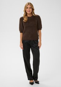 Femme debout portant un pull à manches courtes couleur brun foncé et en tissu duveteux, un pantalon en satin noir à jambes droites, et des talons noirs à bouts pointus avec des nœuds.