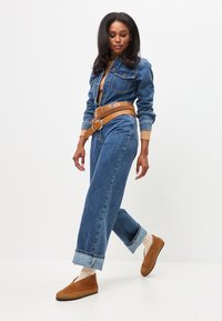 Blauwe denim jumpsuit met een aansluitende taille en brede pijpen, voorzien van een lichtbruine leren riem. Bruine instappers en lichte sokken.