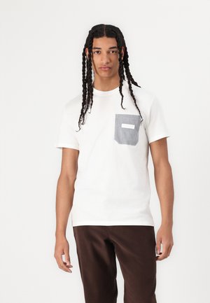 T-shirt en coton blanc avec un détail de poche rayé. Manches courtes et col rond. Associé à un pantalon marron, le modèle se tient face à l'avant.