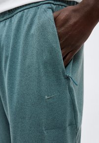 Σκούρο δέρμα χέρι στην τσέπη με φερμουάρ σε τιρκουάζ αθλητικά σορτς Nike με ελαστική μέση, σε λευκό φόντο.