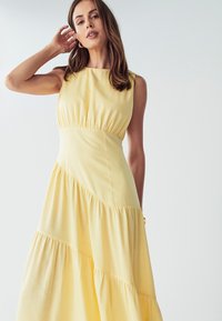 Rumena brezrokavna obleka vsebuje prilegajoč zgornji del, zbrano podrobnost na pasu in večplastno, padajočo krilo z teksturirano površino.