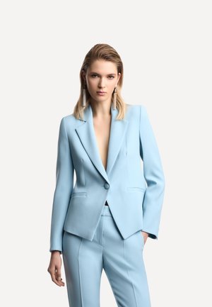 Donna che indossa un completo pantaloni su misura di colore azzurro chiaro con orecchini lunghi, in piedi con una mano in tasca davanti a uno sfondo semplice.