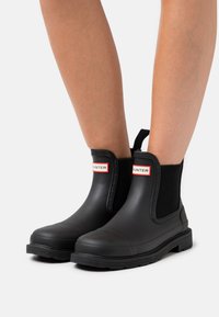 Black rubber ankle boots with elastic side panels, flat soles, and red logo accents on the front. Smooth texture with rounded toe.