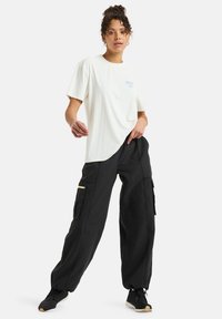 White short-sleeve t-shirt with a blue logo, paired with loose black cargo pants featuring side pockets and yellow zippers.