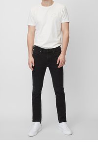 Hombre con camiseta blanca de manga corta, jeans negros ajustados y zapatillas blancas, de pie frente a un fondo sencillo.