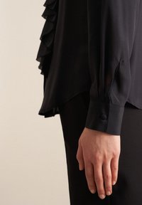 Blusa nera con dettagli arricciati su un lato, maniche lunghe, polsini con bottoni e un tessuto liscio e fluido. Design elegante e sartoriale.