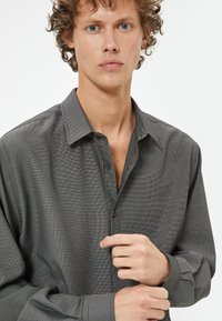 Chemise grise à boutons avec un motif texturé subtil, dotée d'un col et de manches longues. Elle présente une coupe décontractée avec des boutons visibles.