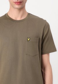 Tricou din bumbac de culoare verde măslin, cu guler rotund, mâneci scurte și un buzunar pe piept, care prezintă un logo galben brodat. Textură netedă.
