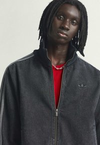 Črna denim jakna z zadrgo in visokim ovratnikom, ki ima dve beli črti na rokavih in Adidasov logotip na prsih. Oblečena je nad rdečo majico.