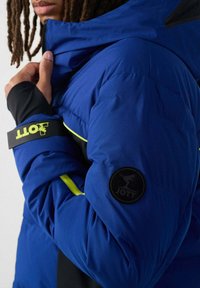 Person mit Dreadlocks, die eine blaue gesteppte Jacke trägt, teilweise geöffnet, mit JOTT-Logo auf dem Ärmel und gelben Akzenten.
