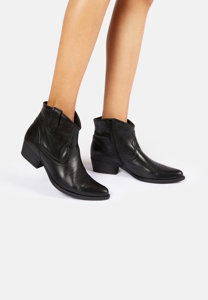 Botas de tobillo de cuero negro con punta afilada, cremallera lateral y tacón bajo apilado. Detalles sutiles texturizados en la parte delantera.