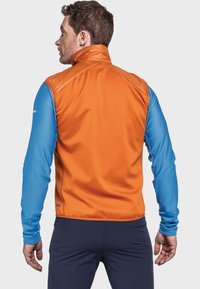 Der farbblockierte Weste hat einen orangefarbenen Rücken und lange blaue Ärmel, gefertigt aus strukturierten, flexiblen Materialien; Zipper sind nicht sichtbar.