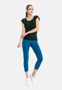 Donkergroene fitted top met kapmouwtjes en een ronde hals, gecombineerd met blauwe cropped leggings en witte sportschoenen.