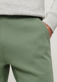 Pantaloni jogger verdi in cotone con una texture liscia, dotati di tasche laterali e una vita aderente, abbinati a un maglione grigio chiaro.
