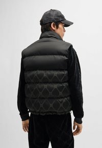 Black padded vest with quilted pattern featuring raised branding; high collar; worn over a black textured outfit; smooth material.