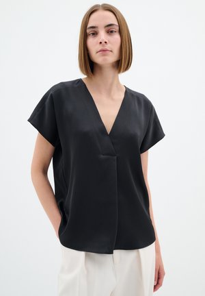 Top corto nero con scollo a V e tessuto testurizzato, caratterizzato da una vestibilità comoda e tasche laterali. Indossato con pantaloni chiari.
