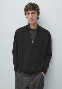 Donkergrijze sweater met rits en geribbelde structuur, voorzien van een hoge kraag en lange mouwen, gedragen over een zwart shirt, gecombineerd met gestructureerde grijze broek.