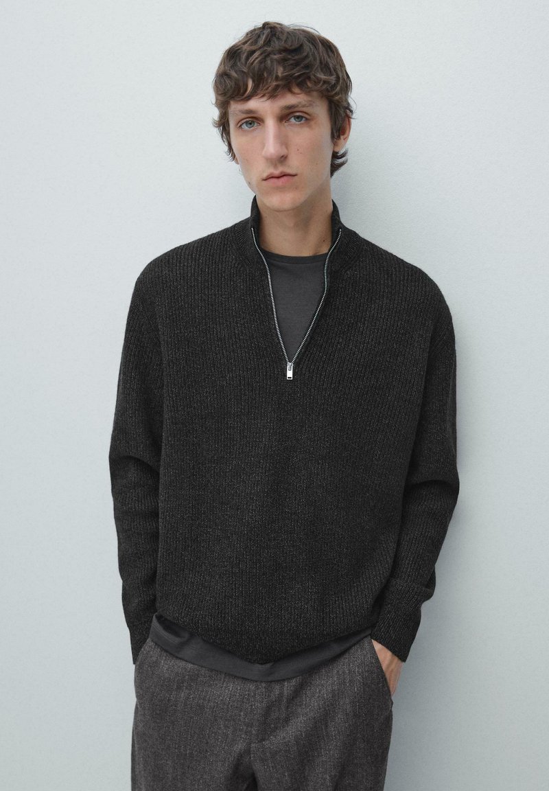 Donkergrijze sweater met rits en geribbelde structuur, voorzien van een hoge kraag en lange mouwen, gedragen over een zwart shirt, gecombineerd met gestructureerde grijze broek.