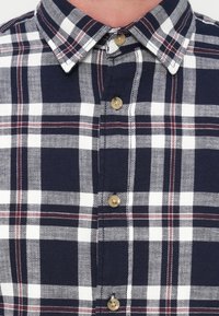 Πουκάμισο με καρώ σχέδιο σε ναυτικό μπλε και λευκό, με γιακά που κουμπώνει κάτω, χρυσά κουμπιά και υφασμάτινη υφή με κόκκινες λεπτομέρειες στο σχέδιο.