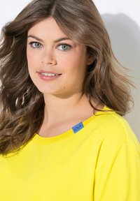 T-shirt jaune vif avec un col rond, des manches courtes et une étiquette bleue sur l'épaule. Tissu doux avec une texture lisse.
