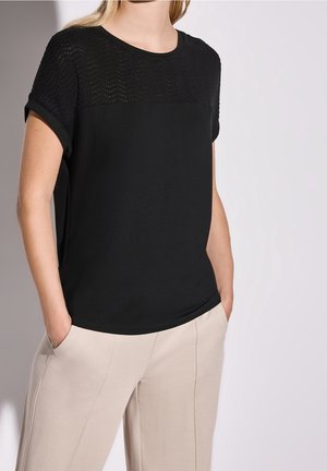 Kobieta ubrana w czarny krótki top z teksturowaną górną częścią i beżowe spodnie, z rękami w kieszeniach, na tle jasnego, gładkiego tła.