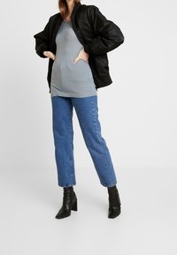 Schwarze Bomberjacke über einem grauen strukturierten Oberteil, kombiniert mit blauen geraden Jeans und schwarzen Stiefeletten mit Absatz. Einfaches, lässiges Outfit.