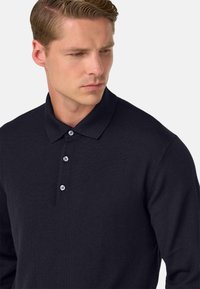 Tums zilas garām piedurknēm polo krekls, izgatavots no teksturēta auduma, ar klasisku apkakli un trīs pogu kabatu ar baltām pogām.