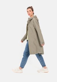 Light olive green waterproof jacket with a hood, zip closure, and snap buttons. Paired with blue jeans and beige sneakers. Long, relaxed fit.