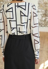 Long-sleeved blouse in sheer white fabric with black geometric patterns, paired with a black high-waisted skirt, highlighting contrasting textures.
