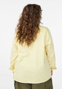 Osoba s dlhými kučeravými hnedými vlasmi, oblečená v bledozltej dlhorukávovej košeli a olivovo zelených cargo nohaviciach, stojaca pred bielym pozadím.