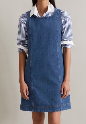 Kobieta w bezrękawnym niebieskim dżinsowym sukience na białej i niebiesko paskowanej koszuli zapinanej na guziki z podwiniętymi rękawami, stojąca na jednolitym tle.