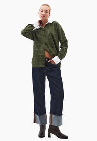 Camicia a quadri verde con colletto bianco, abbinata a jeans blu scuro con risvolti e stivali con tacco marroni.