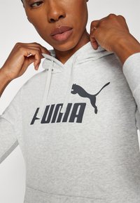 Sivá mikina s kapucňou, s klokaním vreckom, s čiernym potlačeným logom, ktoré znie "PUMA" a so siluetou skáčúceho pumy.