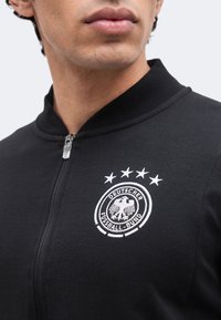 Κοντινό πλάνο ενός ατόμου που φοράει μαύρο μπουφάν με φερμουάρ με το λογότυπο του Deutscher Fußball-Bund και τέσσερα αστέρια κεντημένα στο στήθος.