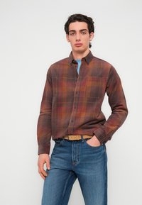 Ανδρικό πουκάμισο με μακριά μανίκια σε καρό μοτίβο με καφέ, κόκκινους και κίτρινους τόνους, με γιακά που κουμπώνει και μία μπροστινή τσέπη, συνδυασμένο με μπλε τζιν.