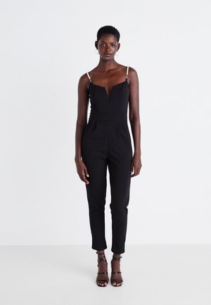 Combinaison noire avec un décolleté en V profond et des bretelles fines ornées de bijoux. Coupe ajustée avec des jambes fuselées. Associée à des talons noirs à lanières.