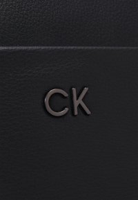 Zwarte leren textuur met een verhoogd "CK"-logo in glanzend metaal. Eenvoudig, afgerond ontwerp met een moderne afwerking.