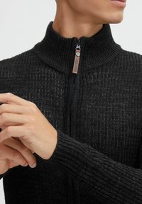 Sort strikket sweater med stående krave, udstyret med lynlåslukning og ribbet tekstur på ærmer og krop. Læderdetalje på lynlåsen.