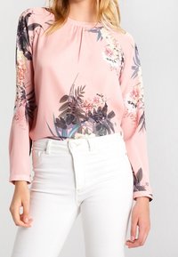 Blusa floreale rosa con maniche lunghe, scollatura arricciata e una texture liscia. Dettagli di foglie verdi e motivi floreali più scuri.