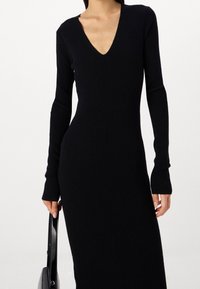 Svart ribbad långärmad klänning med V-ringning, figursydd design och en sidoklyvning. Åtföljs av en svart handväska.