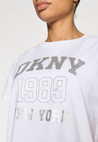 Λευκό βαμβακερό T-shirt με ασημένια τυπωμένα γράμματα: «DKNY 1989» και «NEW YORK», με κλασικό στρογγυλό λαιμό και κοντά μανίκια.