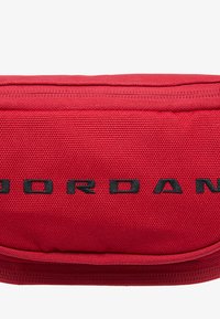 Κόκκινο υφασμάτινο πουγκί με υφή στην επιφάνεια, με εμφανές μαύρο λογότυπο "JORDAN". Κλείσιμο με φερμουάρ στην επάνω άκρη.