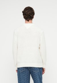 Weißer Strickpullover mit v-förmigem Rücken, geripptem Saum und texturiertem Stoff. Kombiniert mit blauen gemusterten Jeans.