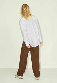White button-up shirt with long sleeves paired with loose brown pants, worn over beige sneakers, viewed from behind against a pale background.