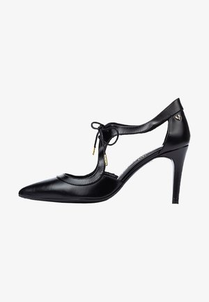 Zapato de tacón alto de cuero negro con punta afilada, lados recortados y detalle de lazo. Presenta acentos dorados y un tacón delgado.