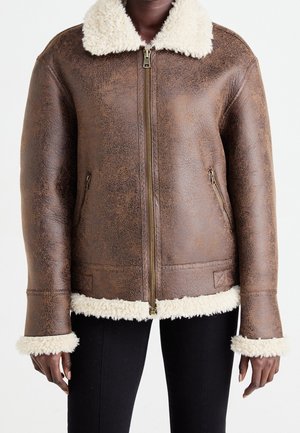 Veste en cuir marron avec doublure en shearling blanc sur le col, les poignets et l'ourlet portée par une personne à la peau foncée et portant un pantalon noir.