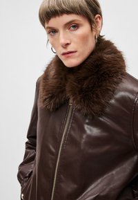 Brun læderjakke med et stort, blødt brunt pelskrave. Har en frontlynlås og sidelommer, glat tekstur og figursyet silhuet.