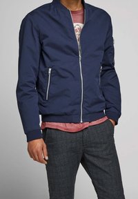 Granatowa kurtka bomberka z kołnierzem stójkowym, zamek z przodu oraz dwiema bocznymi kieszeniami. Miękki materiał z prążkowanymi mankietami i u dołu, nałożona na czerwoną koszulę.