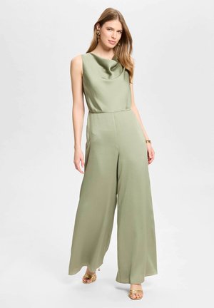 Frau trägt einen ärmellosen, hellgrünen Jumpsuit mit weitem Bein und drapiertem Ausschnitt, steht vor einem schlichten hellen Hintergrund.