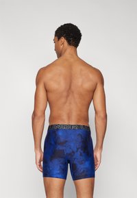 Boxer shorts bleus avec un motif texturé, dotés d'une ceinture noire avec le nom de la marque "UNDR". Visibles de l'arrière sur un torse nu.
